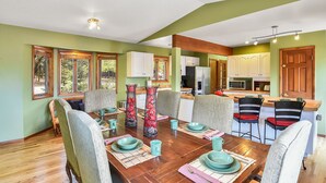 Un comedor con una mesa de madera dispuesta para cuatro personas, rodeada de sillas. Se ve una cocina al fondo con armarios blancos y un refrigerador.