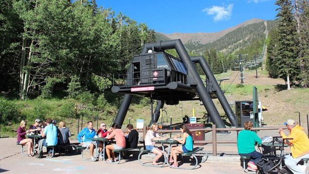 Un grupo de personas sentadas en mesas de picnic al aire libre, con una gran estructura mecánica al fondo.