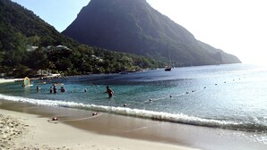 A beach with clear water, a mountain in the background, and a few boats.