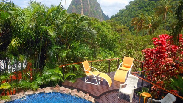 A pool area with lounge chairs and a wooden deck surrounded by lush greenery and a mountain view.