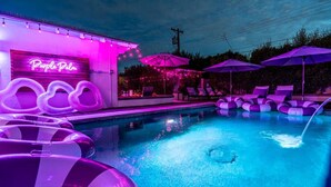 Ein Poolbereich mit Liegestühlen und aufblasbaren Ringen, einer Neonreklame und einem Gebäude mit sichtbarem Eingang.