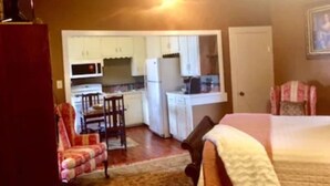 A compact room with a bed, a small desk, a chair, and a kitchenette. - Mary Kate Doyle Suite (Granbury)
