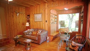 A cozy cabin interior with wooden walls, a leather sofa, a rocking chair, and a dining area with a table and chairs.