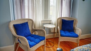 Dos sillas de mimbre con cojines azules, una mesita redonda y un radiador cerca de una ventana con cortinas traslúcidas.