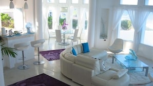 Een moderne woonkamer met een witte bank, een glazen salontafel en een paars vloerkleed. Op de achtergrond bevindt zich een eethoek met witte stoelen en een tafel.