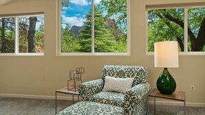 Ein Zimmer mit einem gemusterten Sessel und Hocker, einem kleinen Tisch mit Lampe sowie großen Fenstern mit Blick auf Bäume und Berge.