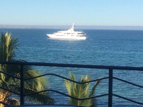 Uno yacht bianco sull'oceano, visto da un balcone con ringhiera e palme sullo sfondo.