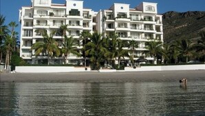 A multi-story hotel with balconies, located near a beach with palm trees and a mountain in the background. - ONLY TRUE BEACH FRONT Condo in REAL La Paz!  Full Sunset View, 2 Pools, Pool Bar (La Paz)