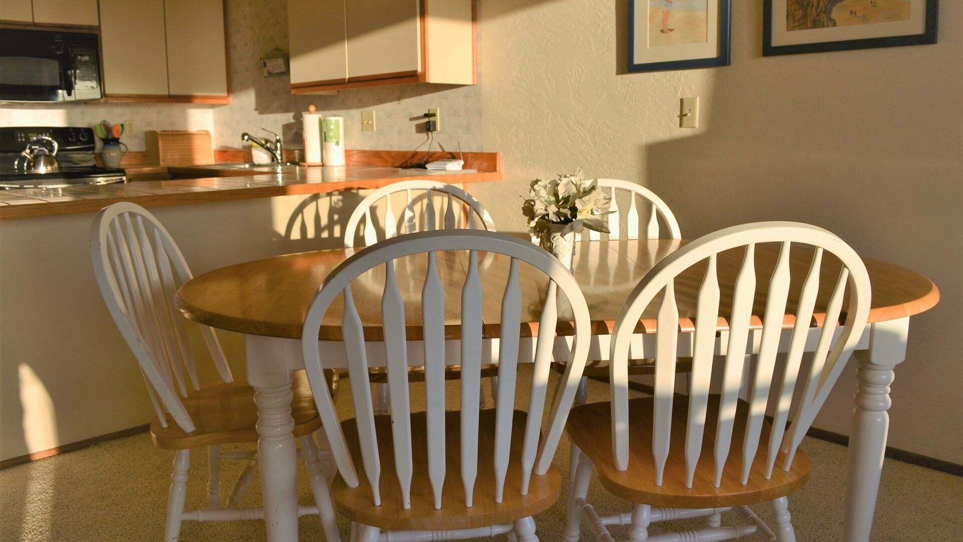 A dining area with a wooden table and white chairs, a kitchen in the background, and a framed picture on the wall.