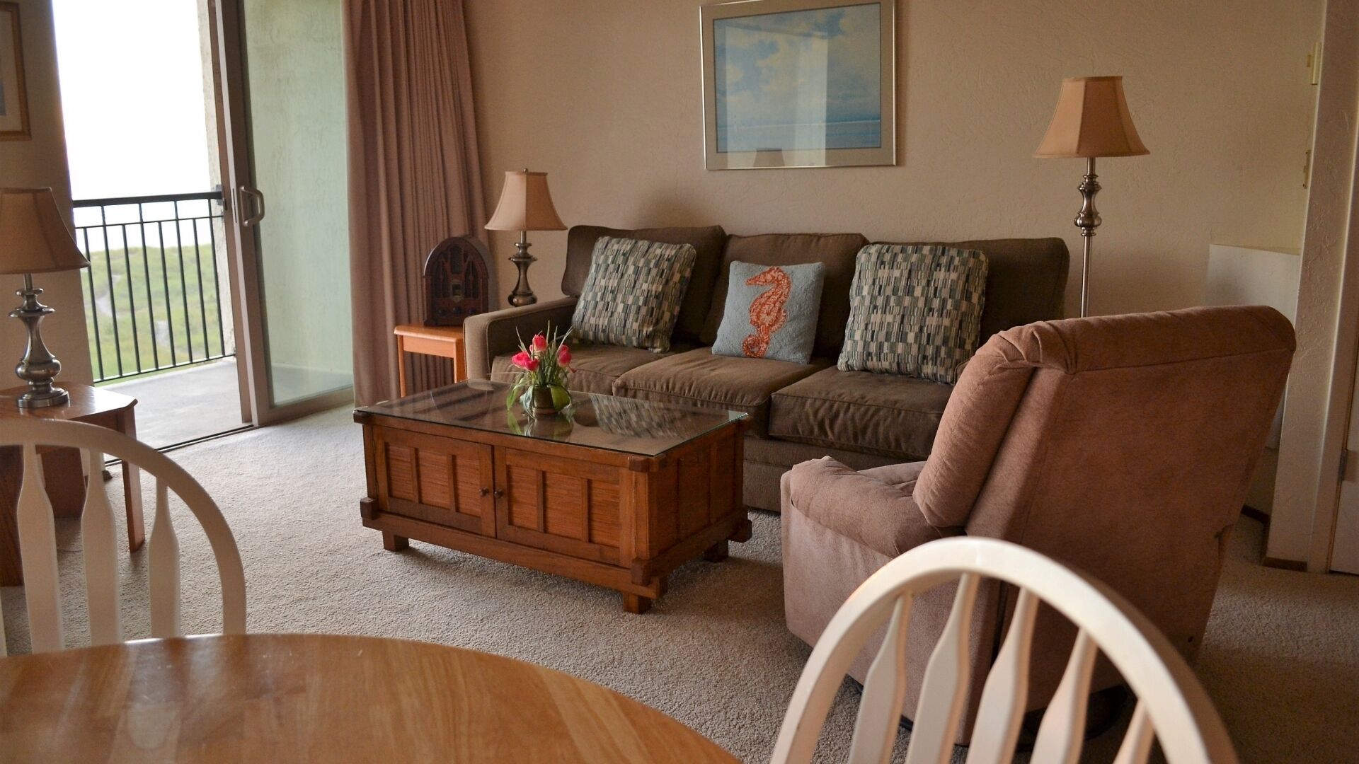 A living room with a sofa, armchairs, a coffee table, and lamps. There is a window with curtains and a view of the outdoors.