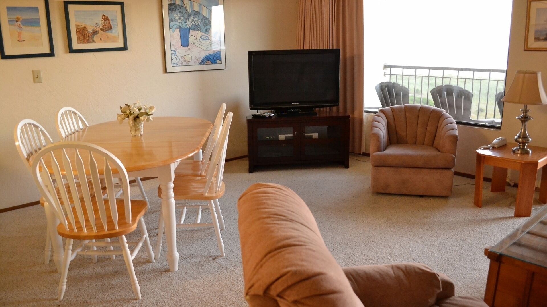 A living room with a round wooden table, white chairs, a brown sofa, a television on a cabinet, and framed artwork on the wall.