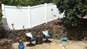 An outdoor seating area with wicker chairs and cushions, a stone wall, and a white fence in the background. - Ocean Views In Paradise (Kailua-Kona)