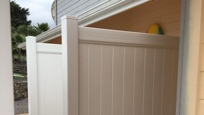 A white wooden fence with vertical paneling, a concrete ground, and a building with a sloped roof in the background. - Ocean Views In Paradise (Kailua-Kona)
