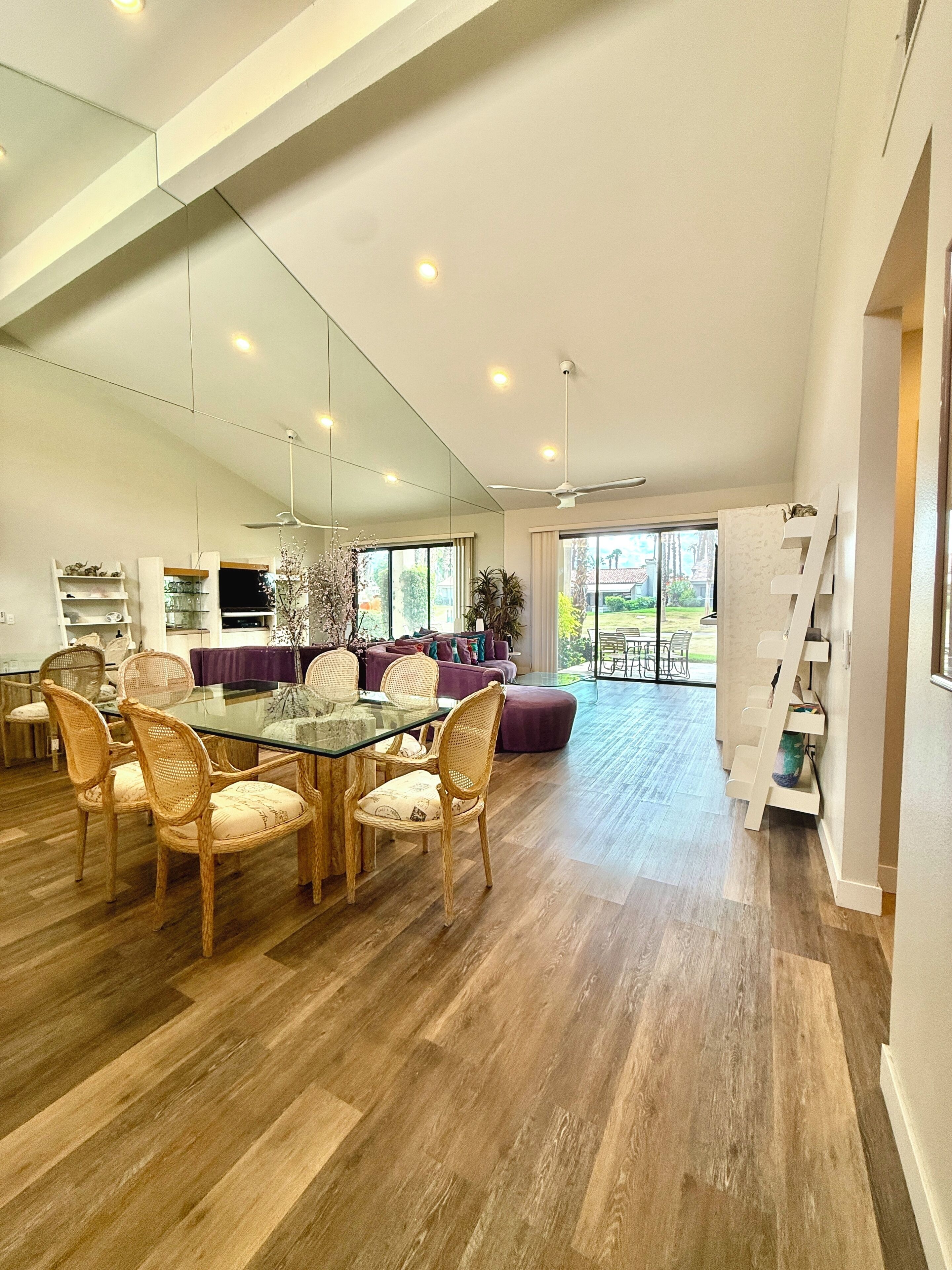A spacious living area with a glass dining table, wicker chairs, a purple sofa, and a large window offering a view of the outdoors.