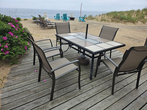 A wooden deck with a table and chairs, overlooking a beach with chairs and a view of the ocean. - Upscale Cottage On The Atlantic Ocean, Feet in Sand (Newbury)