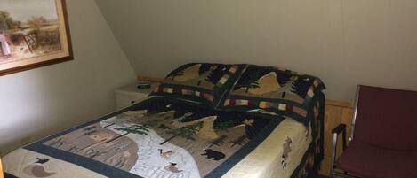 Un letto singolo con copriletto raffigurante scene di fauna selvatica e natura, un comodino con una sveglia e una sedia rossa in una stanza dalle pareti spoglie.
