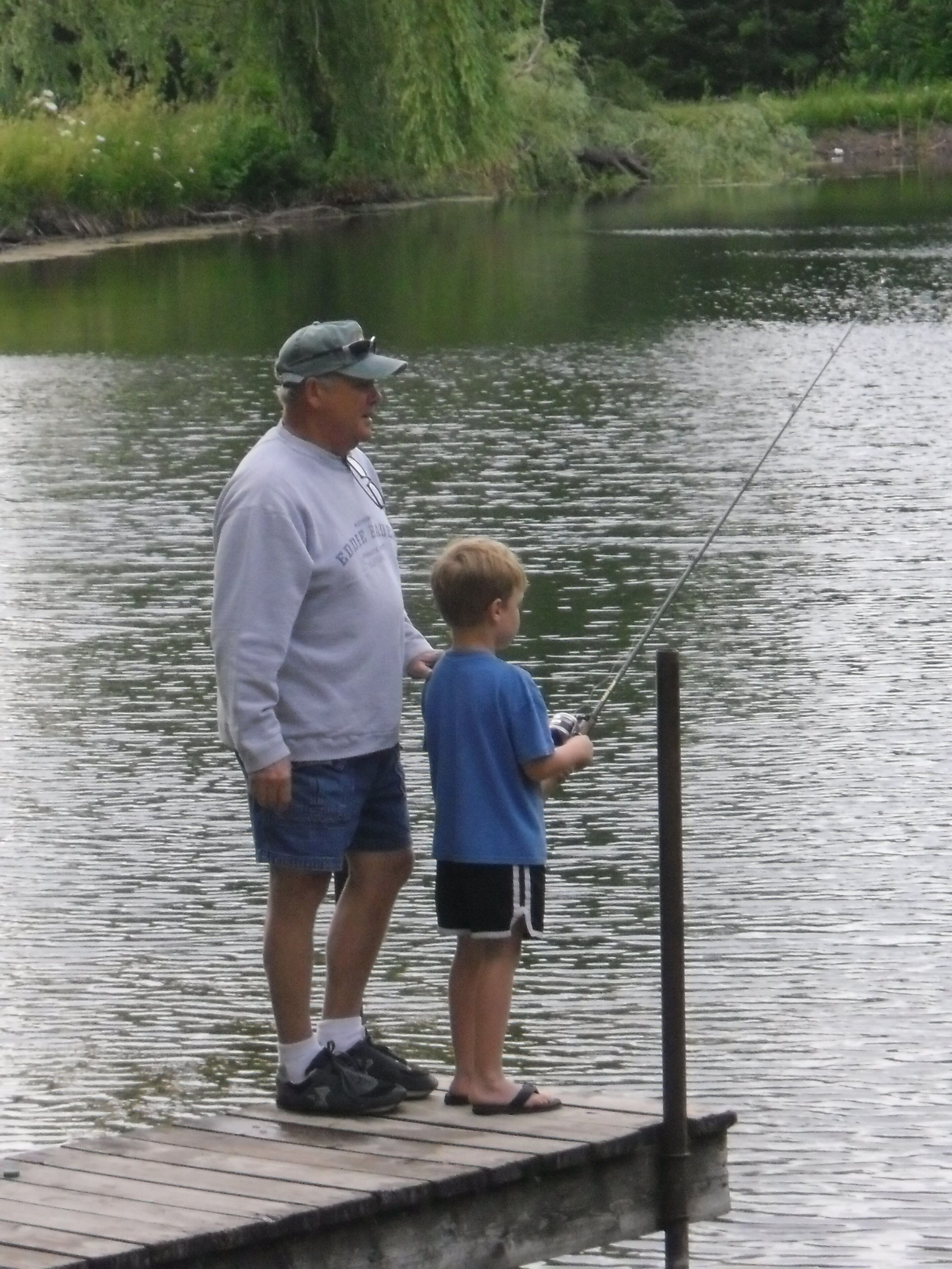 A man and a boy fishing on a wooden dock by a calm lake.