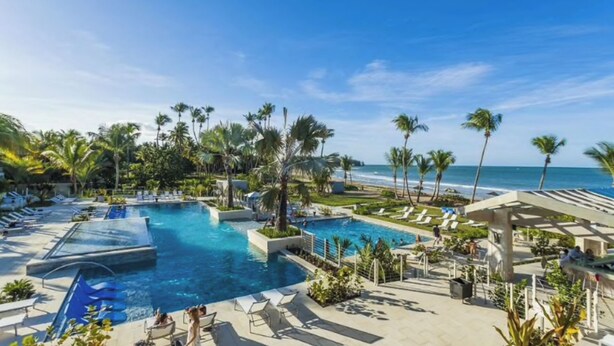Zona de piscina en un resort con varias piscinas, palmeras y vista a la playa.