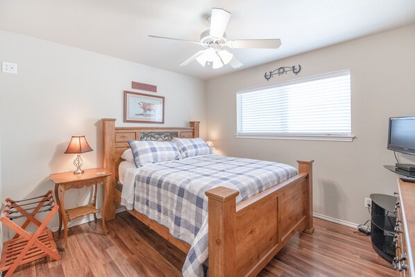 A bedroom with a wooden bed, a ceiling fan, a window with blinds, a TV, and a small table with a lamp.