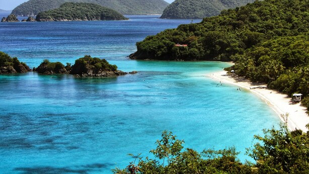 Eine Küstenlandschaft mit klarem türkisblauem Wasser, einem Sandstrand und sanften, grünen Hügeln im Hintergrund.