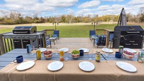 An outdoor dining area with a table set for a meal, surrounded by a wooden deck and a view of a grassy field. - Large beautiful Farmhouse in the heart of PEC - 20 minutes to Sandbanks (Prince Edward)