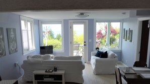 Ett vardagsrum med en soffa, ett soffbord, en tv och ett matbord. Det finns fönster med utsikt över grönområden och en dörr som leder ut.