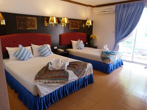 A hotel room with two beds, each with a red headboard, white linens, and blue bed skirts. There are wall-mounted lamps, a nightstand with a phone, and a window with curtains. - Won Won Place (Krabi)
