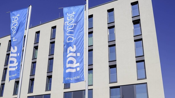 A modern building with multiple windows and three blue flags with white text. - Ibis Budget Konstanz (Konstanz)