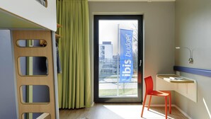 A modern hotel room with a bed, a desk, a chair, a window with a view of buildings, and a curtain. - Ibis Budget Konstanz (Konstanz)