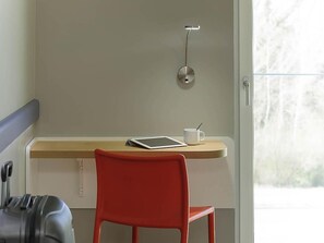 A room with a wooden desk, a red chair, a tablet, a white mug, and a suitcase. - Ibis Budget Konstanz (Konstanz)