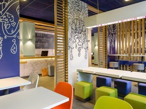 A modern interior with white tables and colorful stools, a wooden slatted partition, and wall art. - Ibis Budget Konstanz (Konstanz)