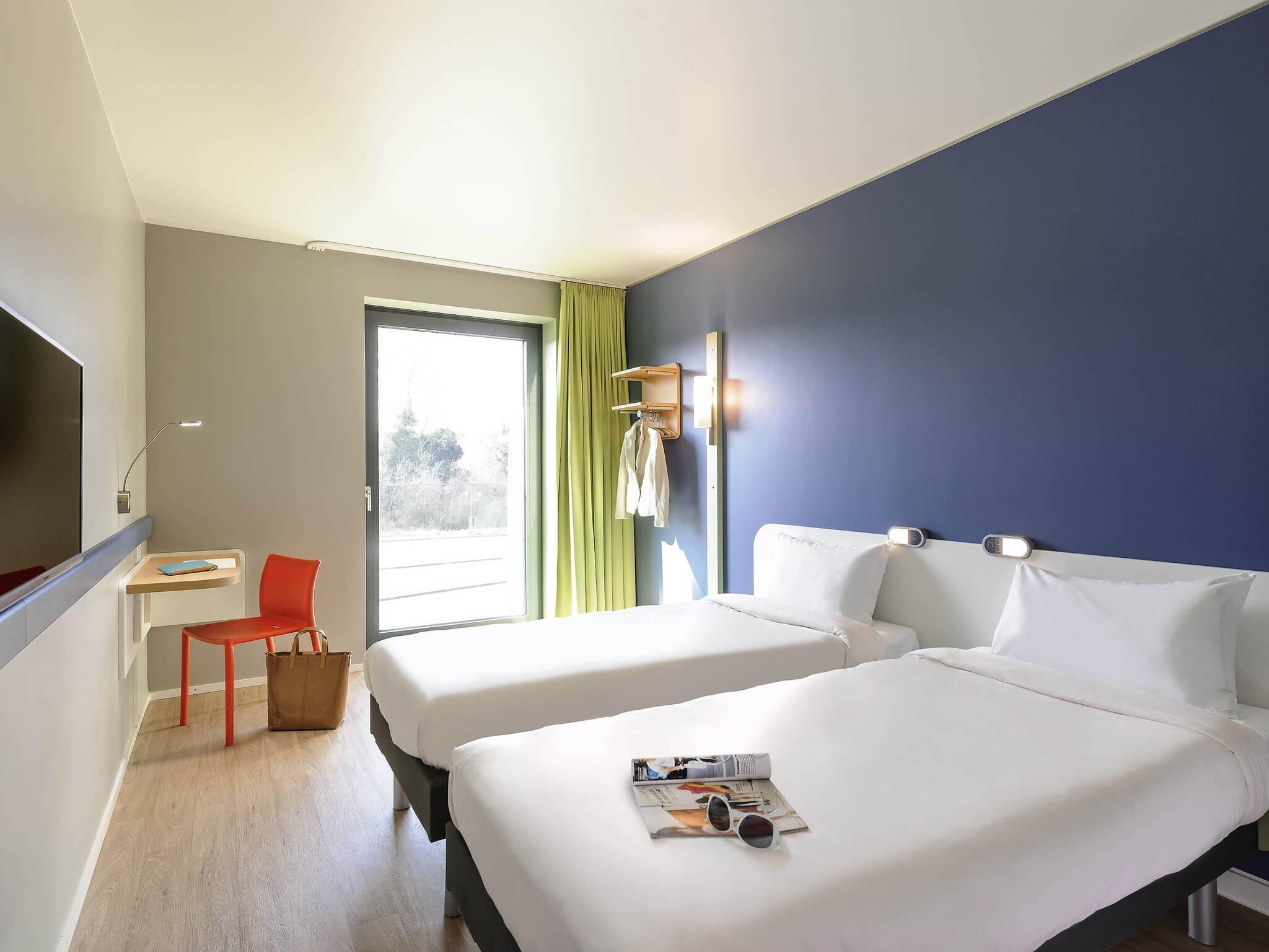 A hotel room with two beds, a desk, a chair, a TV, and a window with curtains.