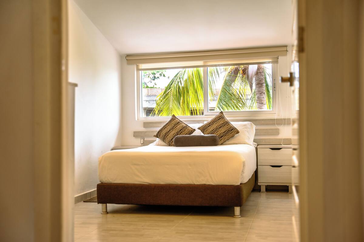 Ein Schlafzimmer mit einem Bett, Kissen, einem Nachttisch und einem Fenster mit Blick auf Palmen.