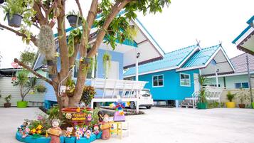 A tree with hanging planters and a bench in front of a blue house with a white car parked nearby.