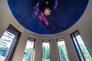 Une pièce dont le plafond est décoré d’un ciel étoilé, avec une étoile centrale qui émet une lumière douce, et qui offre une vue sur les arbres à travers de grandes fenêtres.