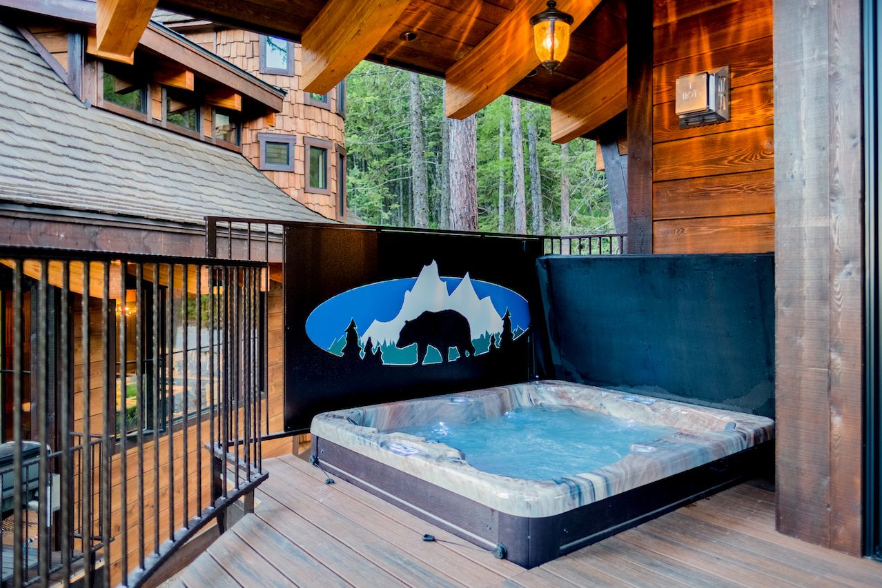 Um deck de madeira com uma banheira de hidromassagem, uma cabana de madeira ao fundo e um mural com o contorno de uma montanha.