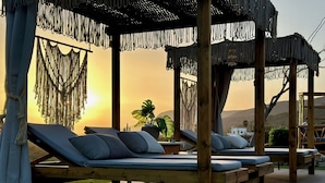 A sunlit outdoor lounge area with wooden loungers, a pergola with macrame fringe, and a view of the sunset over hills. - 9 beds, 7 wc, sauna, jacuzzi, pool, cinema, bar, bbq, firepit, helipad, parking (Chania)