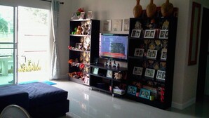 A living room with a TV, shelves with decorative items, and a view of the outdoors. - Sandy Home Sattahip (Sattahip)