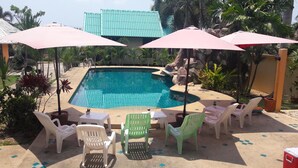 A swimming pool with a tiled deck, surrounded by plastic chairs and umbrellas, and a building with a green roof in the background. - Sandy Home Sattahip (Sattahip)