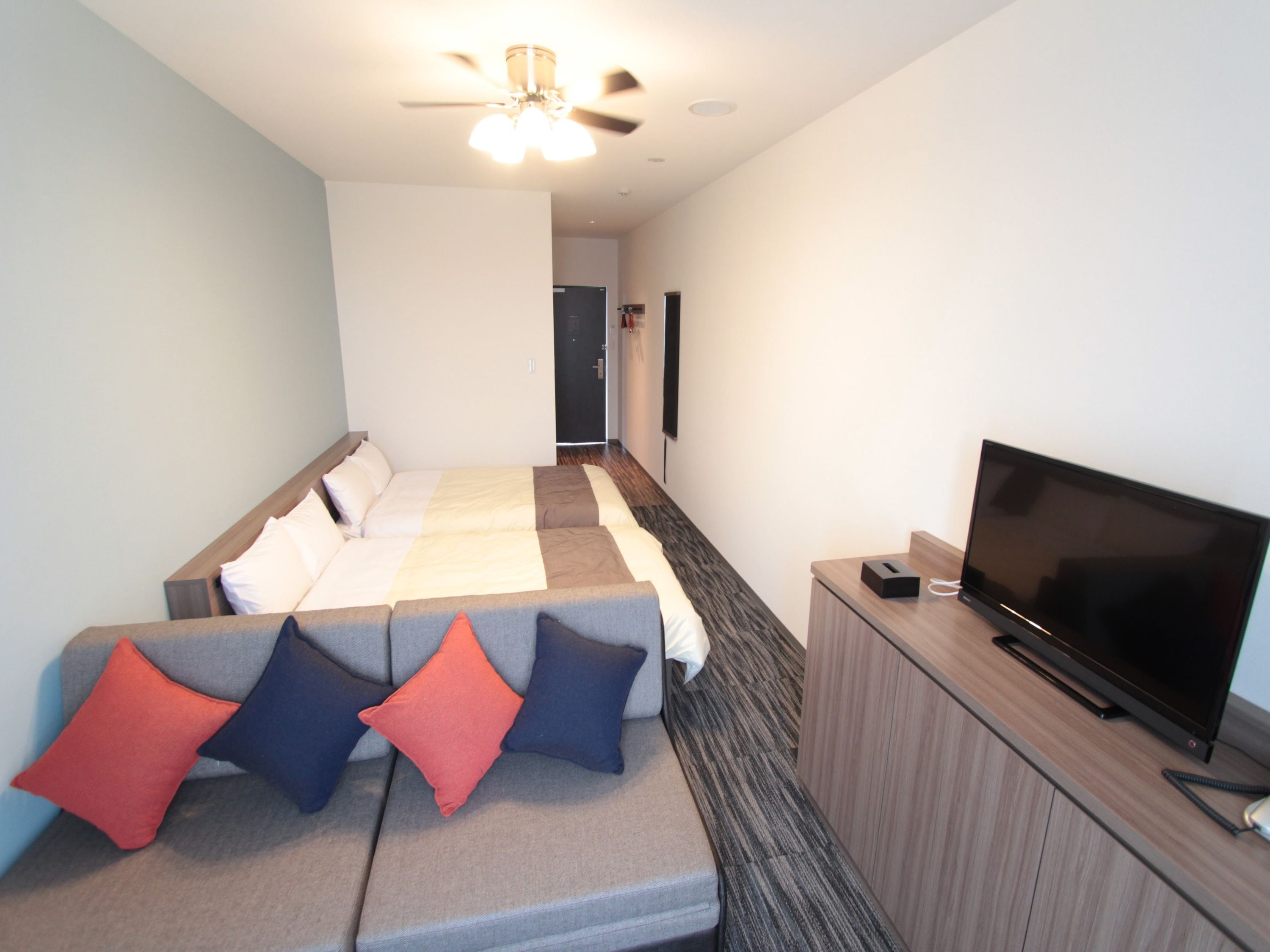 A hotel room with a bed, a sofa with cushions, a television, and a ceiling fan.