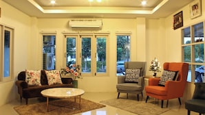 A living room with a brown sofa, a round coffee table, a patterned armchair, and a black armchair. There are three windows with blinds, a ceiling fan, and an air conditioning unit. - Green Apple Villa (Sattahip)