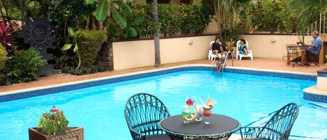 Piscina com mesa e cadeiras, rodeada por vegetação exuberante e um edifício ao fundo.