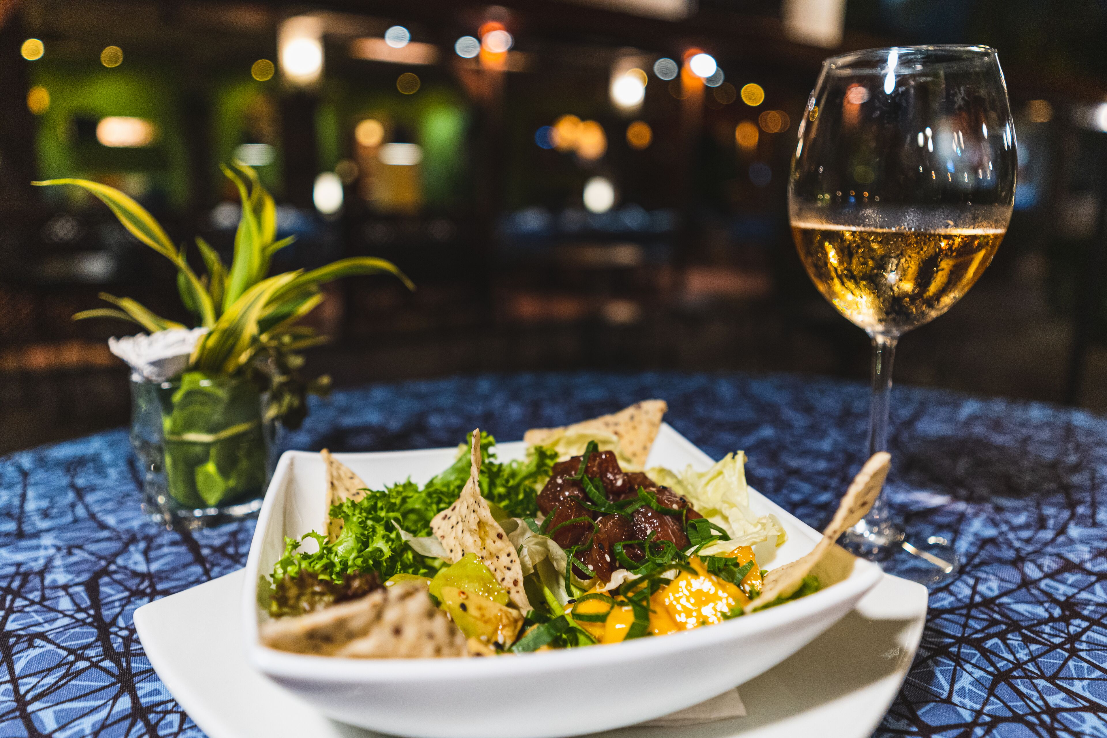 En un plato, una ensalada con una copa de vino blanco sobre una mesa con mantel estampado azul.