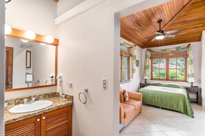 Une salle de bain avec un lavabo, un miroir et un meuble-lavabo, et une chambre avec un lit, une fenêtre et un ventilateur de plafond.