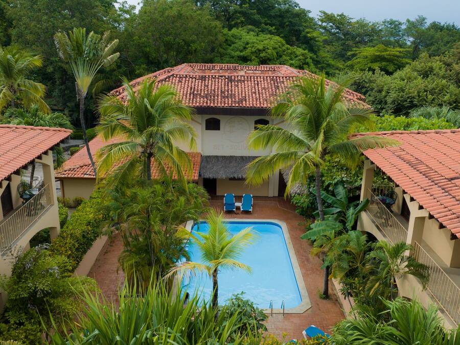 Un resort con una piscina central rodeada de palmeras y un edificio con techo de paja.