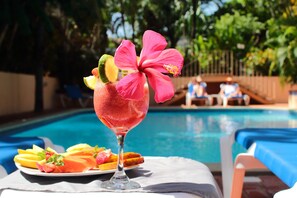 A poolside setting with a drink garnished with fruit and a hibiscus flower, a plate of sliced fruits, and a pool in the background.