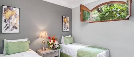 Habitación de hotel con dos camas, una mesita de noche con lámpara, una ventana con vista a áreas verdes y dos cuadros en la pared.