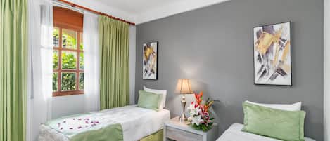 A hotel room with two beds, a nightstand with a lamp and flowers, and two abstract paintings on the walls.
