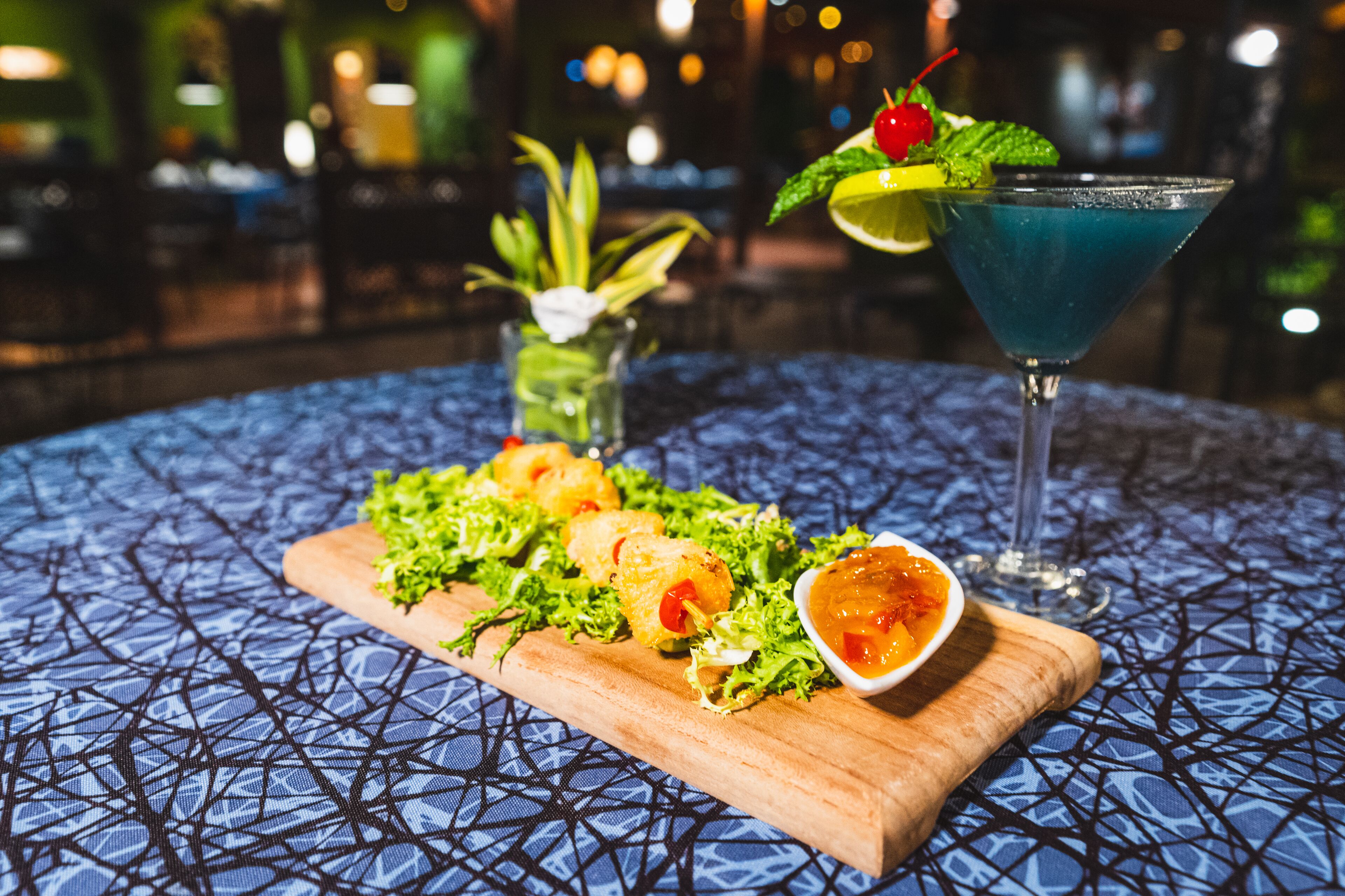 A table with a blue patterned tablecloth, a wooden board with appetizers, a cocktail glass with a blue drink, and a small vase with greenery.