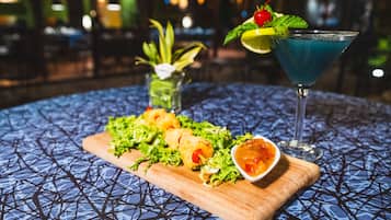 A table with a blue patterned tablecloth, a wooden board with appetizers, a cocktail glass with a blue drink, and a small vase with greenery.